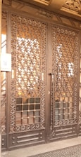A large wrought iron gate with intricate floral and geometric patterns, featuring golden accents. The structure is set against a solid backdrop and has a keypad entry system on the left side.