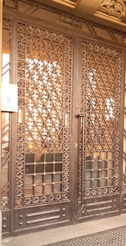 A large wrought iron gate with intricate floral and geometric patterns, featuring golden accents. The structure is set against a solid backdrop and has a keypad entry system on the left side.