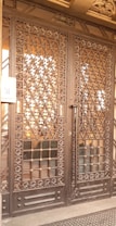 A large wrought iron gate with intricate floral and geometric patterns, featuring golden accents. The structure is set against a solid backdrop and has a keypad entry system on the left side.