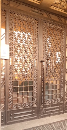 A large wrought iron gate with intricate floral and geometric patterns, featuring golden accents. The structure is set against a solid backdrop and has a keypad entry system on the left side.
