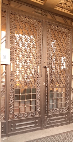 A large wrought iron gate with intricate floral and geometric patterns, featuring golden accents. The structure is set against a solid backdrop and has a keypad entry system on the left side.