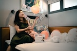 A woman wearing glasses is joyfully playing with an orange balloon in a decorated room. A 'Happy Birthday' banner is hung on the wall, surrounded by balloons and a gift box. There's a patterned pillow and twisted towel sculptures adding to the festive atmosphere.