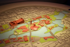 A detailed model of a city layout featuring various buildings and structures in red, surrounded by green areas and water bodies. The model is intricately crafted with several distinctive zones and landmarks, indicating urban planning and landscape design.