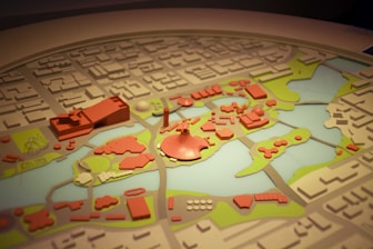 A detailed model of a city layout featuring various buildings and structures in red, surrounded by green areas and water bodies. The model is intricately crafted with several distinctive zones and landmarks, indicating urban planning and landscape design.