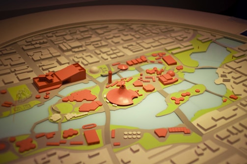 A detailed model of a city layout featuring various buildings and structures in red, surrounded by green areas and water bodies. The model is intricately crafted with several distinctive zones and landmarks, indicating urban planning and landscape design.
