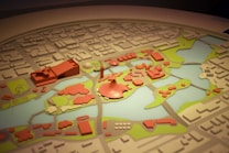 A detailed model of a city layout featuring various buildings and structures in red, surrounded by green areas and water bodies. The model is intricately crafted with several distinctive zones and landmarks, indicating urban planning and landscape design.