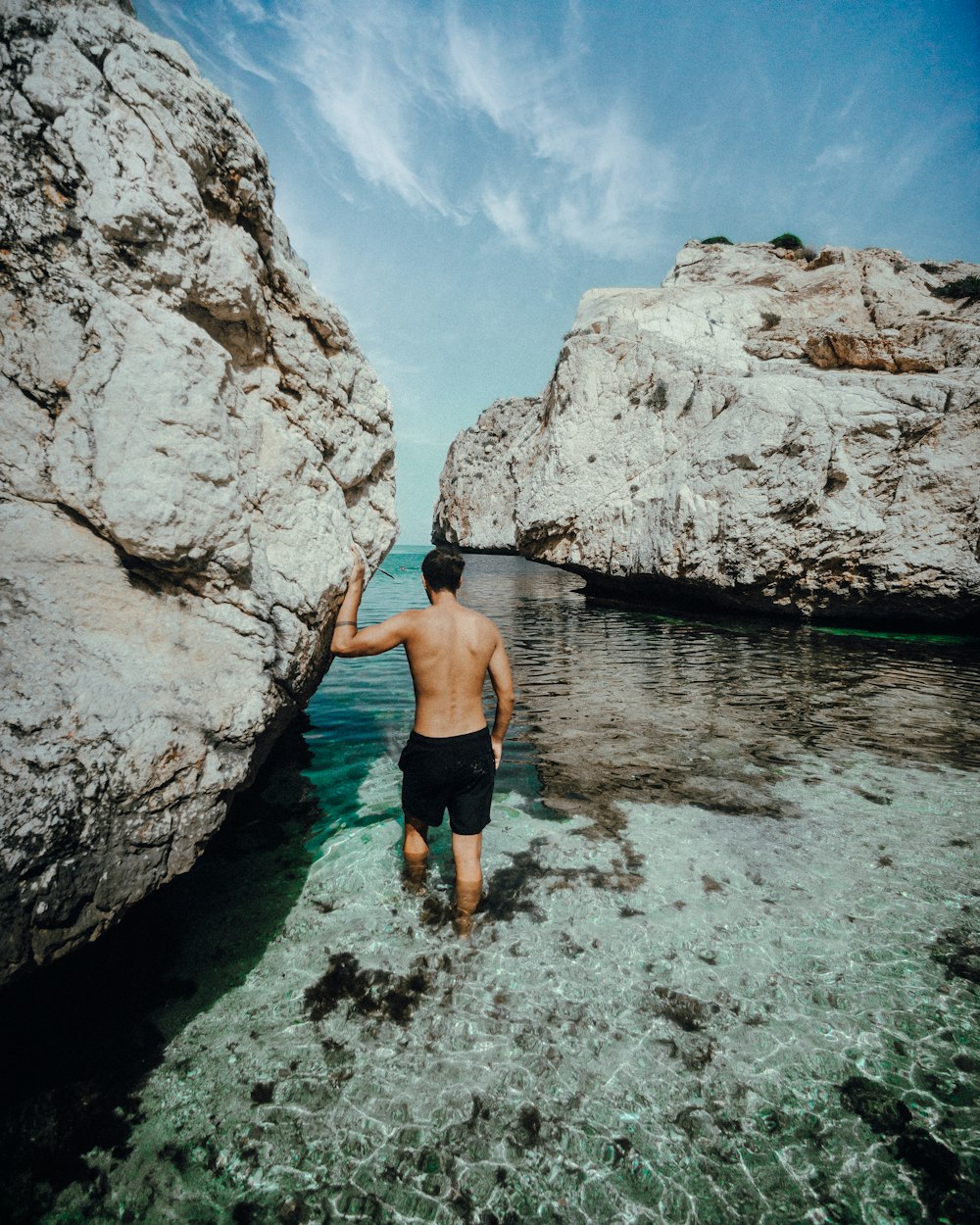 man in blue shorts standing on body of water near brown rock formation during daytime