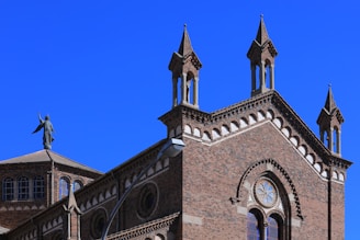 A detailed architectural view of a historical brick building adorned with pointed towers and intricate window designs. A statue of an angel or figure stands atop one of the building's roofs, adding a sense of grandeur.