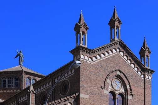 A detailed architectural view of a historical brick building adorned with pointed towers and intricate window designs. A statue of an angel or figure stands atop one of the building's roofs, adding a sense of grandeur.