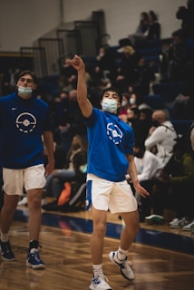 Two basketball players wearing blue shirts and white shorts, both wearing face masks, are on a basketball court. One player is in the middle of a shooting motion, with spectators blurred in the background.