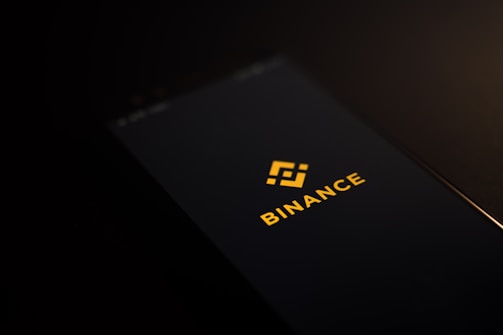 A smartphone screen displaying the Binance logo with yellow text and an icon in the center against a dark background.