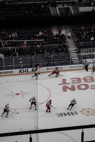 Hockey players on the ice rink are actively engaged in a game, with some wearing red jerseys and others in white. The arena appears to be a professional setting with numerous spectators in the stands, some of whom are standing and others seated. The rink has various advertisements and markings on the ice.