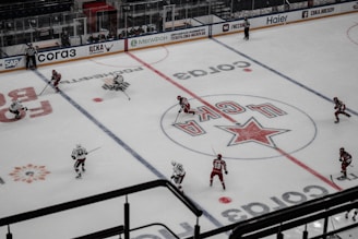 An ice hockey game is in progress on a rink with players wearing red and white uniforms competing against each other. The ice is marked with logos and lines, and two referees are visible. The ice rink has advertising boards around the perimeter, and there are empty seats in the stands.