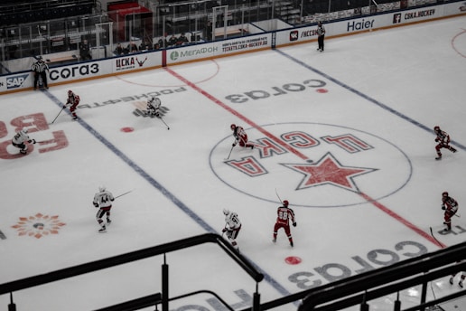 An ice hockey game is in progress on a rink with players wearing red and white uniforms competing against each other. The ice is marked with logos and lines, and two referees are visible. The ice rink has advertising boards around the perimeter, and there are empty seats in the stands.