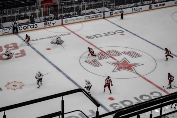 An ice hockey game is in progress on a rink with players wearing red and white uniforms competing against each other. The ice is marked with logos and lines, and two referees are visible. The ice rink has advertising boards around the perimeter, and there are empty seats in the stands.