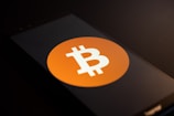 A smartphone screen displays the Bitcoin logo, a white 'B' with two vertical lines, set against an orange circle. The phone is placed on a dark surface with low lighting, casting shadows across the image.