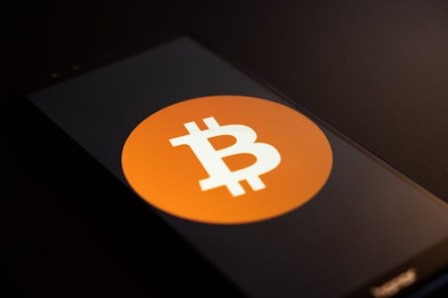 A smartphone screen displays the Bitcoin logo, a white 'B' with two vertical lines, set against an orange circle. The phone is placed on a dark surface with low lighting, casting shadows across the image.