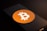 A smartphone screen displays the Bitcoin logo, a white 'B' with two vertical lines, set against an orange circle. The phone is placed on a dark surface with low lighting, casting shadows across the image.