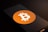 A smartphone screen displays the Bitcoin logo, a white 'B' with two vertical lines, set against an orange circle. The phone is placed on a dark surface with low lighting, casting shadows across the image.