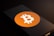 A smartphone screen displays the Bitcoin logo, a white 'B' with two vertical lines, set against an orange circle. The phone is placed on a dark surface with low lighting, casting shadows across the image.
