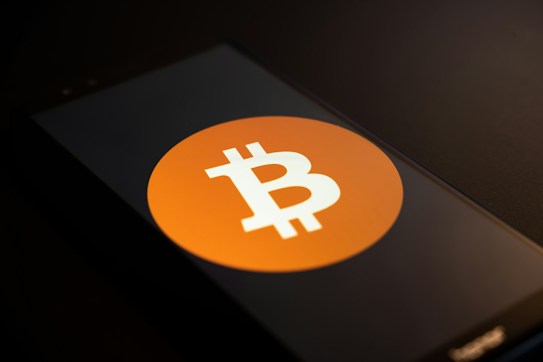 A smartphone screen displays the Bitcoin logo, a white 'B' with two vertical lines, set against an orange circle. The phone is placed on a dark surface with low lighting, casting shadows across the image.