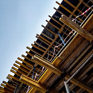 A partially constructed building structure with wooden beams, columns, and supporting elements. Red and white caution tape is visible, likely indicating areas under construction or restricted access. The clear blue sky serves as the backdrop.