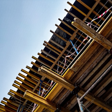A partially constructed building structure with wooden beams, columns, and supporting elements. Red and white caution tape is visible, likely indicating areas under construction or restricted access. The clear blue sky serves as the backdrop.