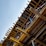 A partially constructed building structure with wooden beams, columns, and supporting elements. Red and white caution tape is visible, likely indicating areas under construction or restricted access. The clear blue sky serves as the backdrop.