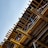 A partially constructed building structure with wooden beams, columns, and supporting elements. Red and white caution tape is visible, likely indicating areas under construction or restricted access. The clear blue sky serves as the backdrop.