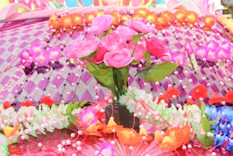 A colorful arrangement of artificial flowers sits on a decorated surface. Pink roses, surrounded by multicolored floral garlands and accents, create a vibrant and festive atmosphere. The background consists of a pattern of pinkish-purple checks with additional flowers and small ornaments.
