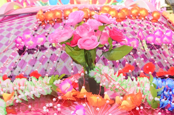 A colorful arrangement of artificial flowers sits on a decorated surface. Pink roses, surrounded by multicolored floral garlands and accents, create a vibrant and festive atmosphere. The background consists of a pattern of pinkish-purple checks with additional flowers and small ornaments.