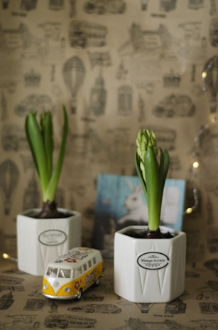 Two white pots labeled 'Vintage Home' contain green budding plants. In front of the pots is a miniature model of a vintage Volkswagen van with colorful decals. The background features a whimsical pattern with illustrations of air balloons, buses, and various other items in a subtle sepia tone.