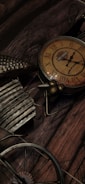 A vintage-looking clock with Roman numerals and a bicycle design is placed against a wooden surface. The brown and rust tones complement the aged aesthetic, and the clock's face bears a logo.