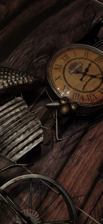 A vintage-looking clock with Roman numerals and a bicycle design is placed against a wooden surface. The brown and rust tones complement the aged aesthetic, and the clock's face bears a logo.