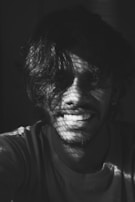 A black and white portrait of a person smiling, with light casting intricate patterns of shadows across their face and hair.