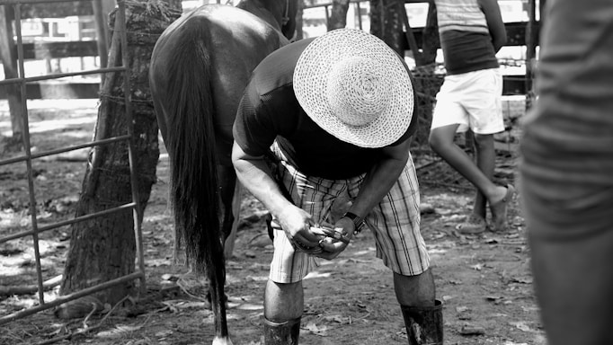 A person wearing a straw hat, plaid shorts, and boots is bending over, possibly grooming the hooves of a horse. Another person stands nearby, leaning against a fence, dressed in casual summer wear. The setting appears to be outdoors, likely at a farm or stable, with a rustic, shaded environment.