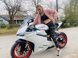 A person in a pink jacket and camo leggings is standing next to a white Ducati 959 Panigale motorcycle with red rims. The setting is outdoors, on a gray road, with trees and a blue house in the background.