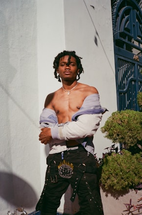 A person with dreadlocks stands confidently with arms crossed. They are wearing black pants with decorative chains and a partially opened jacket. The background includes a white wall and a blue metal gate, along with some greenery to the side.