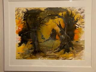A framed abstract painting features a vivid display of colors and shapes, with a predominant use of dark and earthy tones. Silhouetted tree forms and branches create a mysterious, woodland scene. The background displays a blend of oranges and yellows, evoking a sense of warmth and depth.