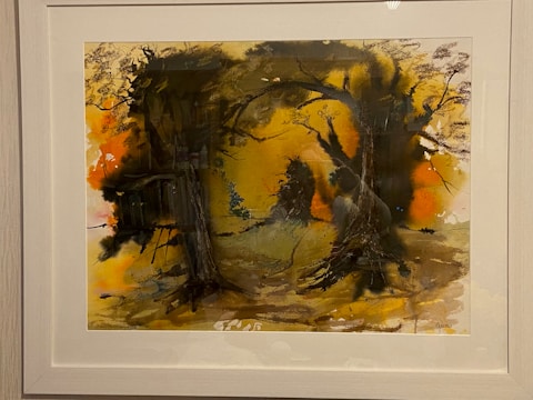 A framed abstract painting features a vivid display of colors and shapes, with a predominant use of dark and earthy tones. Silhouetted tree forms and branches create a mysterious, woodland scene. The background displays a blend of oranges and yellows, evoking a sense of warmth and depth.