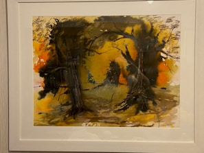A framed abstract painting features a vivid display of colors and shapes, with a predominant use of dark and earthy tones. Silhouetted tree forms and branches create a mysterious, woodland scene. The background displays a blend of oranges and yellows, evoking a sense of warmth and depth.