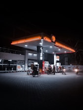 A gas station at night is illuminated by overhead lights, with a Shell logo prominently displayed. Several motorcycles and riders are gathered near the pumps, each under the artificial glow. The surrounding area is dimly lit, emphasizing the bright canopy of the station.