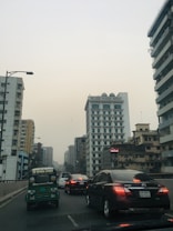 A cityscape with multiple tall buildings lining both sides of a road. Several cars, including a green autorickshaw, are visible, and the scene appears to be during daylight with an overcast or hazy sky. The buildings vary in architectural style, indicating an urban setting.