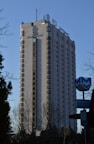 A tall hotel building with numerous windows and a logo labeled 'Hotel Marinela' near the top. In the foreground, there are bare trees and a sign with a subway symbol and street name written in both Cyrillic and English. The sky is clear and blue, suggesting it is daytime.
