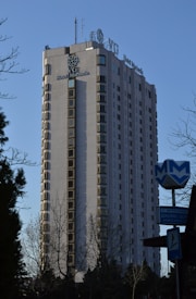 A tall hotel building with numerous windows and a logo labeled 'Hotel Marinela' near the top. In the foreground, there are bare trees and a sign with a subway symbol and street name written in both Cyrillic and English. The sky is clear and blue, suggesting it is daytime.