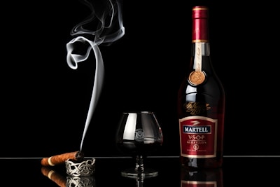 A bottle of Martell VSOP Medaillon cognac stands alongside a glass filled with the dark liquid. To the left of the glass, a lit cigar rests on an ornate ashtray, with a swirl of smoke rising elegantly into the air. The background is completely black, emphasizing the rich colors and reflective surface beneath the objects.
