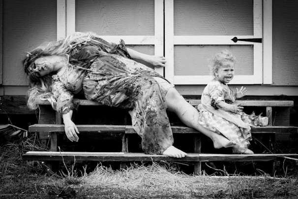 A black and white image depicts an adult woman and a small child on a set of wooden steps. The woman, in a patterned dress, appears unconscious or in a deep sleep with her hair draped over her face. The child, also in a dress, is covered in dirt and is visibly upset, sitting with an open mouth as if crying. The setting is outdoors, near a wooden structure.