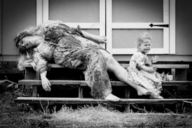 A black and white image depicts an adult woman and a small child on a set of wooden steps. The woman, in a patterned dress, appears unconscious or in a deep sleep with her hair draped over her face. The child, also in a dress, is covered in dirt and is visibly upset, sitting with an open mouth as if crying. The setting is outdoors, near a wooden structure.