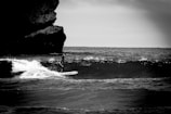 A lone surfer rides a wave near a rocky shoreline, captured in black and white. The ocean appears dynamic with waves breaking, and a large rock formation looms on the left side of the frame.