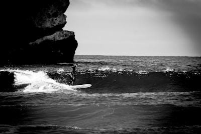 A lone surfer rides a wave near a rocky shoreline, captured in black and white. The ocean appears dynamic with waves breaking, and a large rock formation looms on the left side of the frame.